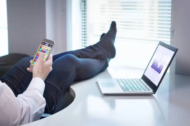 Photo of Lazy office worker playing mobile game with smartphone during work hours. Avoiding his job and being lazy with feet   and socks on table. Useless and relaxing man doing nothing and forget his job.
