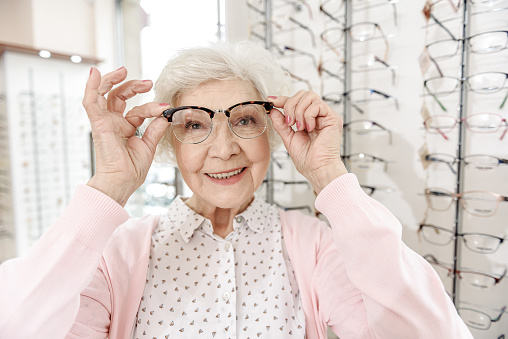 Happy smiling senior lady wearing glasses is looking at camera with interest. She standing near showcase. Portrait