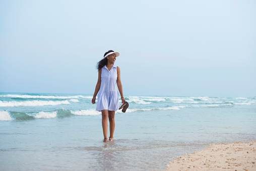 Happy tourist woman walking barefoot on seashore. Woman wearing blue dress and sun visor, holding flip flops in hand.