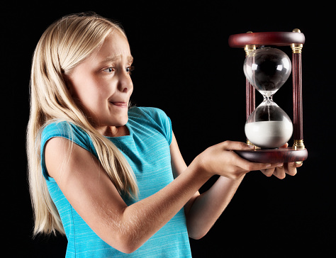 A cute little blonde girl holds a wooden hourglass carefully, looking anxious, possibly worried about time passing or a deadline.