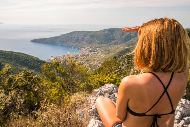Waist up photo of a young woman enjoying the sea breeze while she is relaxing on the top of a hill with a view on the village Komiza. Vis island. Dalmatia. Croatia. Adriatic Sea.