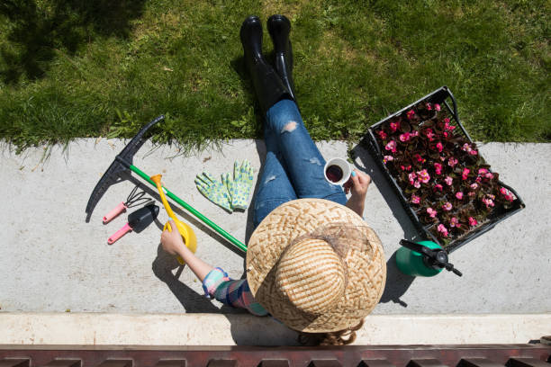 Young woman gardener sitting on the floor in the yard resting and drinking coffee