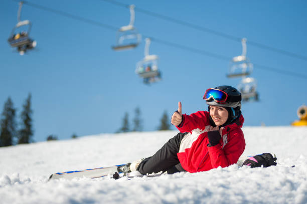 Woman skier with ski at winer resort in sunny day Smiling girl lying with skis on snowy at mountain top, showing thumb up gesture of good class and looking at the camera in sunny day at a winter resort with ski lifts and blue sky in background. Close-up thumbs up for skiing stock pictures, royalty-free photos & images