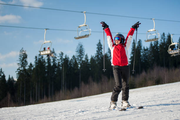 Woman skier with ski at winer resort in sunny day Female skier wearing helmet, red jacket and ski goggles standing on snowy slope with hands raised up in sunny day with forest and blue sky in background. Carpathian, Bukovel bukovel ski resort ukraine stock pictures, royalty-free photos & images