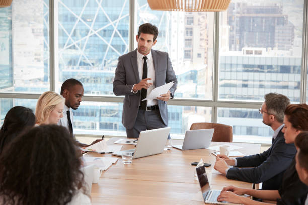 istock Businessman Stands To Address Meeting Around Board Table 677810070