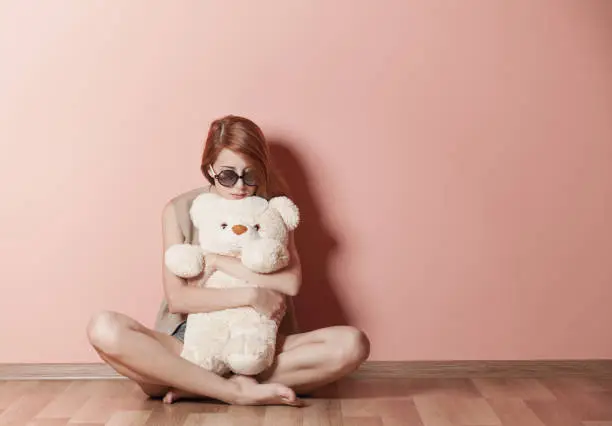 Photo of photo of beautiful young woman hugging teddy bear and sitting near the wall