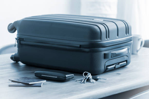 A modern, black, plastic suitcase lying on a table beside a coffee cup, passport, glasses case and keys. The travel case has a plastic carry handle and a textured finish. The selective focus is on the front left corner of the suitcase.