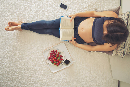 Young pregnant woman, lying in bed with smartphone, book, coffee and fruits, motherhood concept