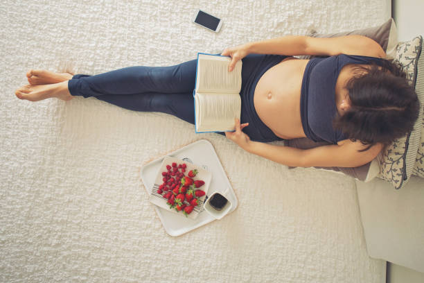 jovem mulher grávida, deitada na cama com o smartphone, livro, café e frutas, conceito de maternidade - blueberry food fruit berry fruit - fotografias e filmes do acervo