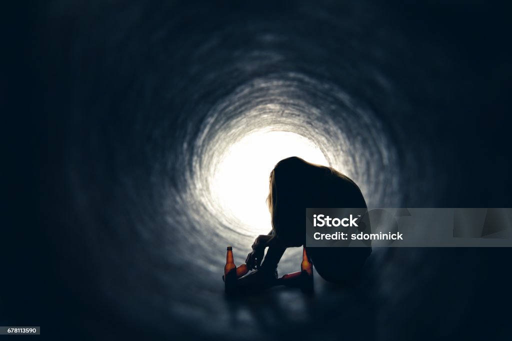 Alcoholic With Empty Beer Bottles In Tunnel A drunk female sitting in a dark tunnel with her head sown surrounded by empty beer bottles. Crying Stock Photo