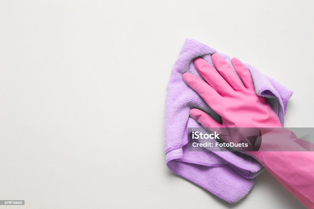 Woman's hand in pink rubber protective glove wiping white wall from dust with dry rag. Early spring cleaning or regular clean up. Maid cleans house. Cleaning Stock Photo