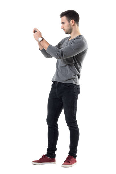 Profile view of serious young casual man holding cellphone taking photo Profile view of serious young casual man holding cellphone taking photo. Full body length portrait isolated over white studio background. canvas shoe photos stock pictures, royalty-free photos & images