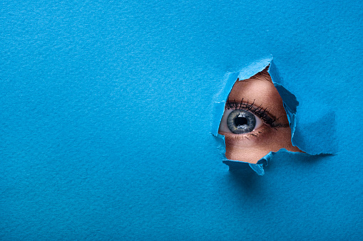 A female eye looks through a hole in a paper blue background.