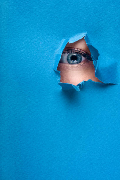 istock A female eye looks through a hole in a paper blue background. 678207300