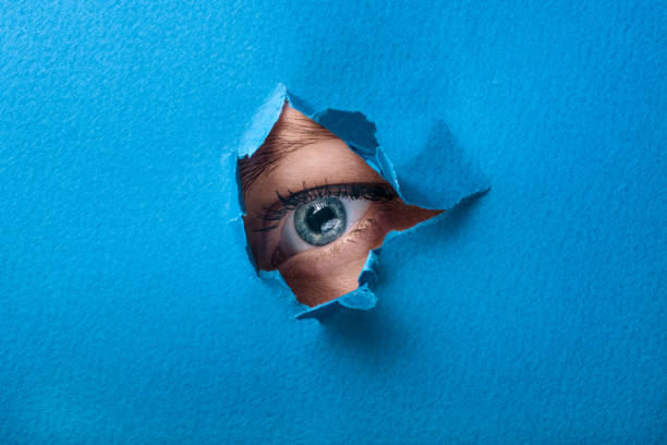 istock A female eye looks through a hole in a paper blue background. 678207304