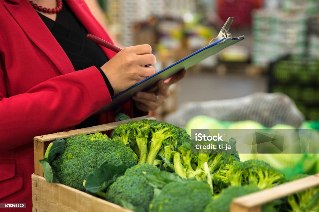 All as on list Woman in a red jacket checking broccoli in a wooden crate at a fruit and vegetable warehouse in Slovenia, Europe. She is holding a clipboard and a pen. Waist down image. Food Stock Photo