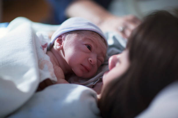 un neonato di razza mista asiatico caucasico in un bambino berretto blu poggia sul petto delle sue madri asiatiche brune e le fissa negli occhi per la prima volta - asian ethnicity baby mother family foto e immagini stock