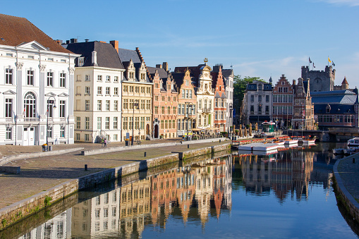View of traditional Belgian buildings next to the Leie's riverside in Ghent. There are just a few people around and the water is reflecting the colorful cityscape.