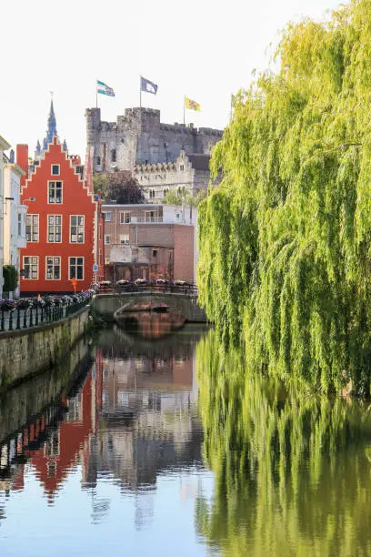 View of the reflection, on the calm waters of the river Leie, of traditional Belgian buildings, the gravensteen castle and some green big trees.