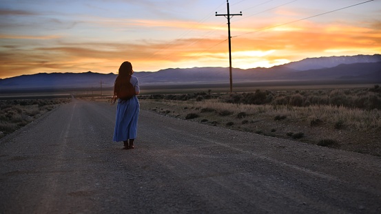 A traditionally dressed girl with dark hair stares out at a desert sunset in Nevada on a dirt road. Amish, western, Native American girl.