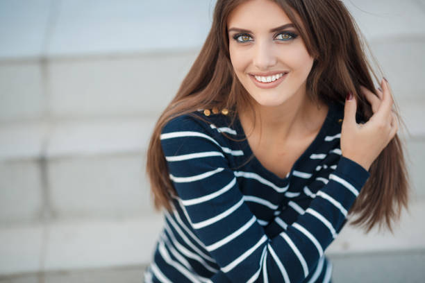 Young beautiful woman brunette with long straight hair and gray-green eyes,a nice smile and white straight teeth,light makeup,dressed in a striped t-shirt and blue jeans,spends time in the city in the spring,sitting on the steps,one in the fresh air
