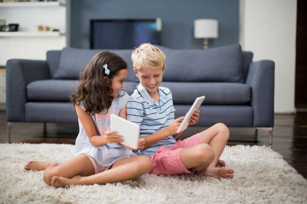 istock Siblings sitting on rug and using digital tablet in living room 678443180