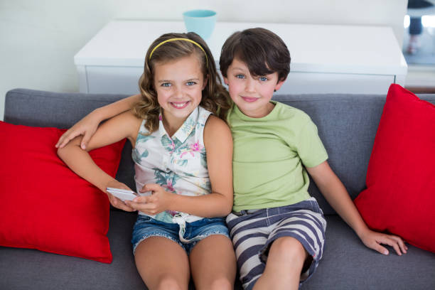 istock Sibling sitting on sofa with mobile phone in living room 678466760