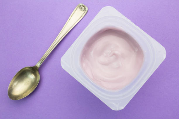yogur con sabor a frutas de fresa sanas con colorante natural en vaso de plástico aislado sobre fondo púrpura con cuchara de plata pequeño - vista superior - yogurt yogurt container strawberry spoon fotografías e imágenes de stock