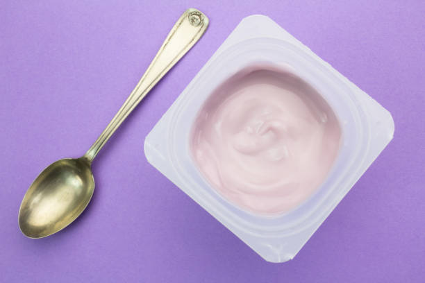 yogurt sano aromatizzato alla frutta alla fragola con colorazione naturale in tazza di plastica isolata su sfondo viola con piccolo cucchiaio d'argento - vista dall'alto - yogurt yogurt container strawberry spoon foto e immagini stock