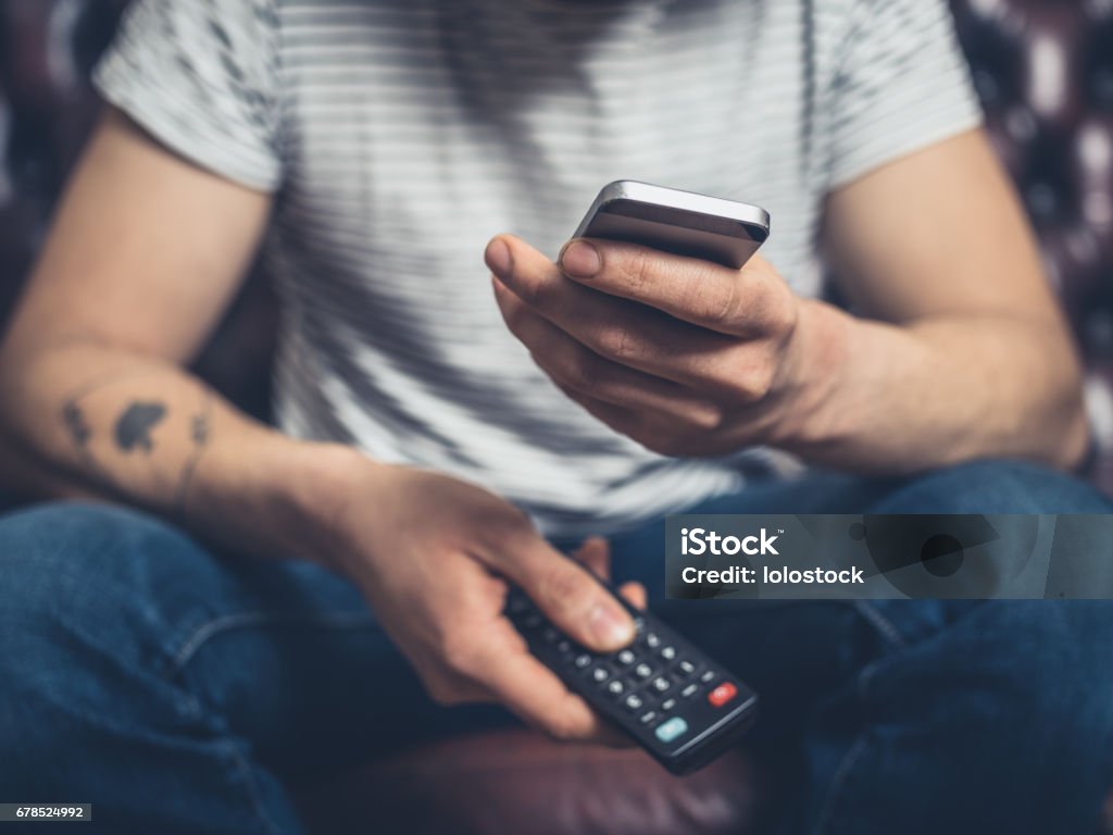 Man on sofa with phone and remote A young man is sitting on a sofa with a remote control and a smartphone Television Set Stock Photo