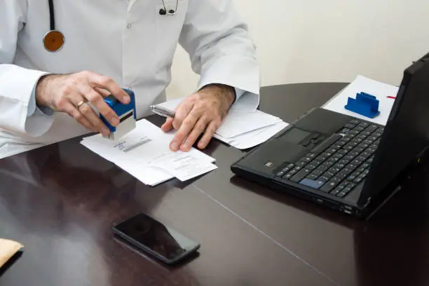 A doctor in a white coat sitting at the table and affix a stamp to the document. Doctor's hand stamped on the prescription. The doctor writes a prescription at his desk. The doctor sits at a table and signed prescription issued.