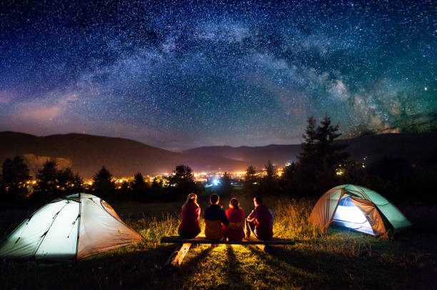 Friends hikers sitting beside camp and tents in the night Friends hikers sitting on a bench made of logs and watching fire together beside camp and tents in the night. On the background beautiful starry sky, mountains and luminous town. Rear view bonfire photos stock pictures, royalty-free photos & images