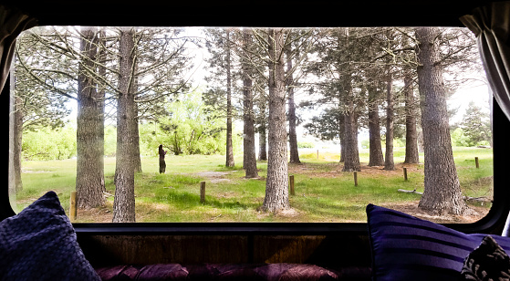 View through the back window of a vintage camper van of a woman standing in the woods, enjoying her early morning coffee.
