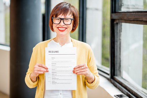 Portrait of an older happy woman holding insurance document standing indoors near the window
