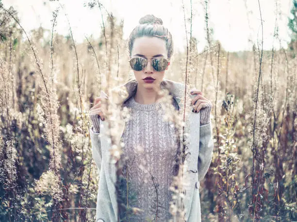 Outdoor fashion photo of young beautiful lady in autumn landscape with dry flowers. Knitted sweater, sunglusses, wine lipstick. Warm Autumn. Warm Spring