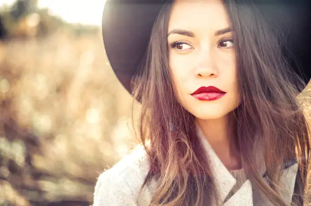 Outdoor fashion photo of young beautiful lady in autumn landscape with dry flowers. Gray coat, black hat, wine lipstick. Warm Autumn. Warm Spring