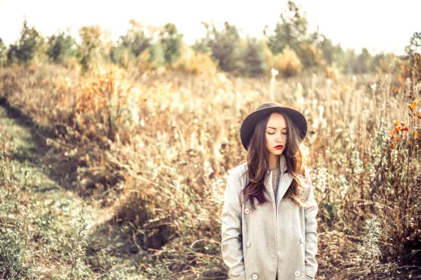 Outdoor fashion photo of young beautiful lady in autumn landscape with dry flowers. Gray coat, black hat, wine lipstick. Warm Autumn. Warm Spring