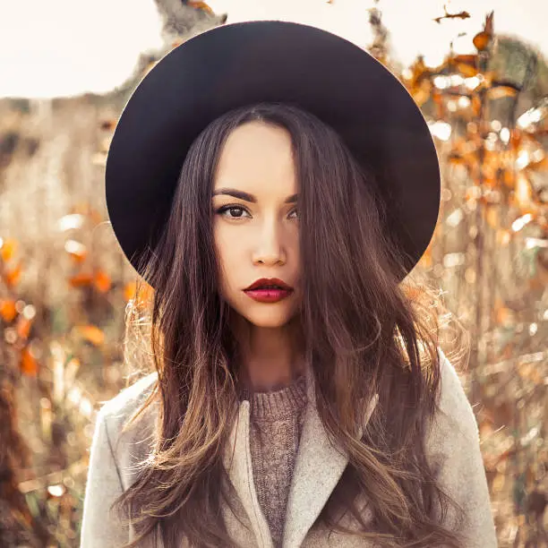 Outdoor fashion photo of young beautiful lady in autumn landscape with dry flowers. Gray coat, black hat, wine lipstick. Warm Autumn. Warm Spring