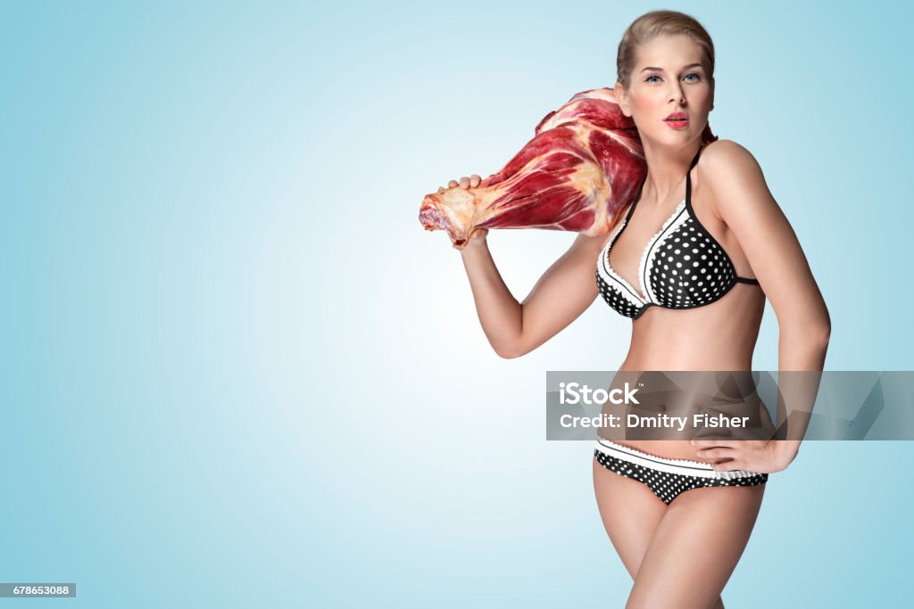 Sexy cook. Hungry. A creative portrait of a beautiful girl in bikini holding raw meat on her shoulder. Bikini Stock Photo