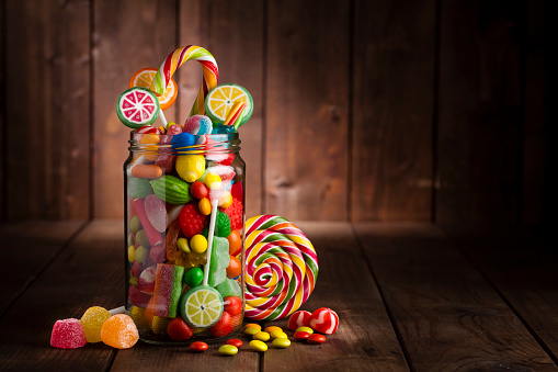 Front view of an open candy jar filled with multi colored candies, lollipops and jelly beans standing on rustic wooden table. Some candies are spilled out of the jar directly on the background. DSRL low key studio photo taken with Canon EOS 5D Mk II and Canon EF 100mm f/2.8L Macro IS USM