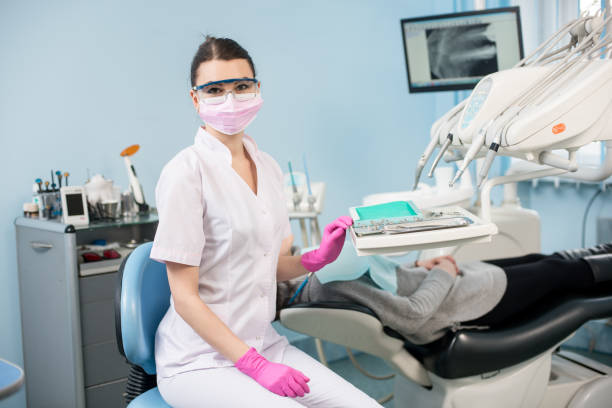 Portrait of attractive female dentist with patient in the dental clinic. Doctor wearing glasses, mask, uniform and pink gloves. On the background monitor with X-ray the patient's teeth. Dentistry Portrait of attractive female dentist with patient in the dental clinic. Doctor wearing glasses, mask, white uniform and pink gloves. On the background monitor with X-ray the patient's teeth. Dentistry Dental Cleanings stock pictures, royalty-free photos & images