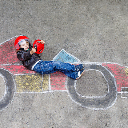 Funny kid boy having fun with race car picture drawing with colorful chalks. Creative leisure for children outdoors in summer
