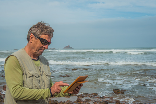 Senior photographer surfing  the net on digital tablet on the red stone Atlantic shore, Legzira, Sidi Ifni Province, Morocco, Africa. Nikon D800.