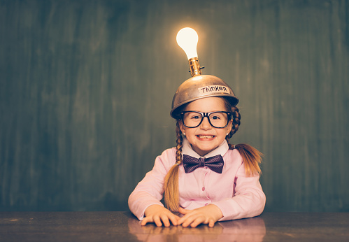 A young female nerd girl sits in a classroom setting with a thinking cap on her head. She has an excited look on her face as the light bulb is turned on and she is getting loads of ideas. She is wearing a pink cardigan and bow tie. Learning is fun when you have ideas.