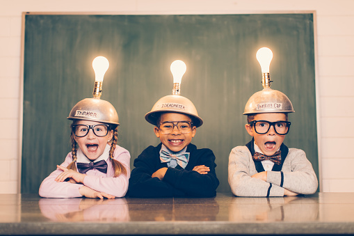 Three young nerds sit in a classroom setting with a thinking cap on their heads. They are suprised and smiling as their light bulbs are lit as the new ideas are flowing. They're wearing cardigans and bow ties. Learning is fun when you have ideas.