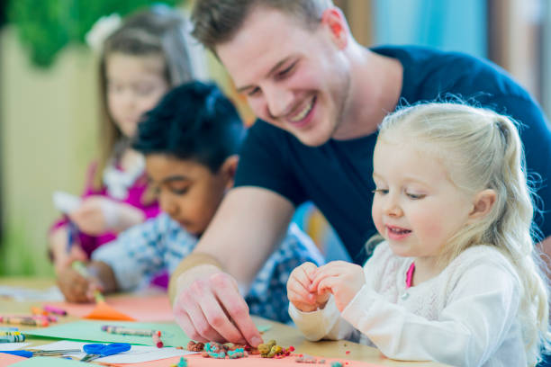 Help From Teacher A multi-ethnic group of young children are indoors at a preschool. They are wearing casual clothing. Their male teacher is helping them color with crayons. A Caucasian girl is smiling in front. preschool classroom jobs stock pictures, royalty-free photos & images