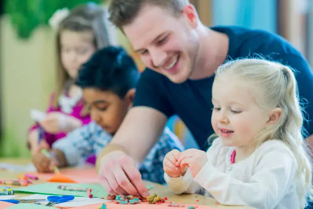 A multi-ethnic group of young children are indoors at a preschool. They are wearing casual clothing. Their male teacher is helping them color with crayons. A Caucasian girl is smiling in front.