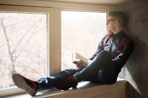 Fashion portrait of a handsome young man sitting by the window and reading a book.