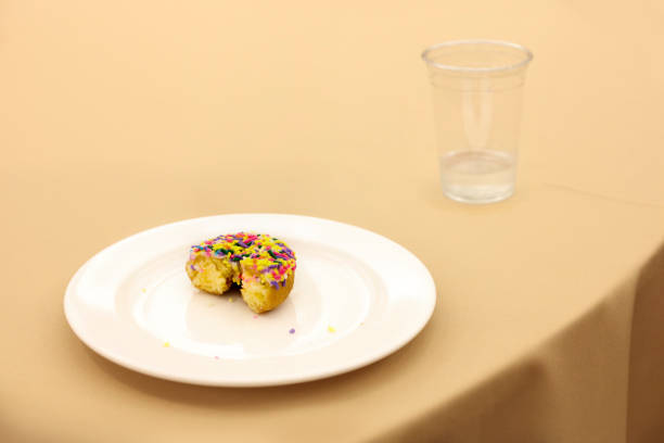 A half-eaten rainbow sprinkles donut left on a plate next to an almost emptied plastic cup.