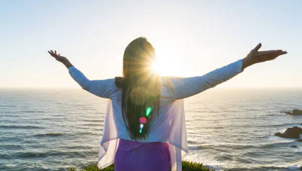 Woman with hands uplifted in worship and adoration against the warm rays of the setting sun overlooking the Pacific Ocean.